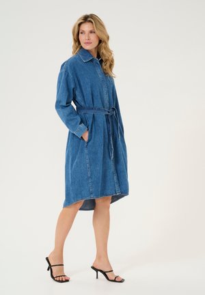 Denimmekko, jossa on kaulus, pitkät hihat ja sidottava vyötärö. Ominaisuuksina sininen väri, taskut ja polven alapuolelle ulottuva, kaareva helma.