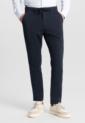 Marineblauwe op maat gemaakte broek met een elastische tailleband, voorkeisjes en een slim fit. Gecombineerd met beige sneakers en een wit shirt.