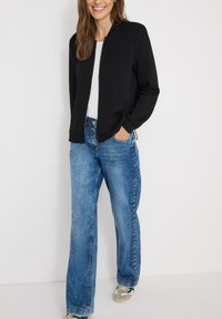 Chaqueta bomber negra con cremallera frontal, camiseta blanca debajo, jeans de pierna ancha de color azul claro y zapatillas con acentos verdes.