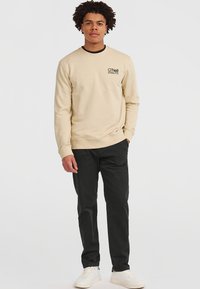 Felpa beige con colletto rotondo, logo ricamato sul petto, polsini e fondo a costine. Abbinata a pantaloni neri e sneaker bianche.