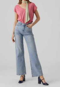 Pink short-sleeve top, light blue wide-leg jeans, and navy block-heel shoes. A green scarf tied around the neck provides an accent.
