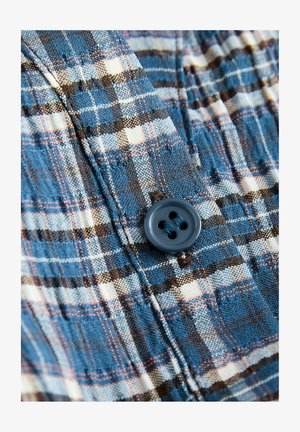 Sininen ruutupaita, jossa on vaaleita ja tummia ruutuja, tummansinisellä neljän napin, tekstuurilla varustetulla kankaalla, jossa on näkyviä ompelutietoja.