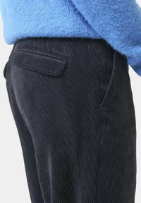 Schwarze Cordhosen mit vertikaler Rippstruktur, ausgestattet mit seitlichen Taschen und einer Paspeltasche auf der Rückseite, kombiniert mit einem flauschigen blauen Pullover.