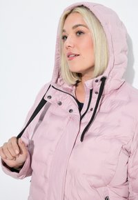 Kvinna med blond bob som bär en ljusrosa pufferjacka med huva, som håller i ett svart snöre, mot en vit bakgrund.