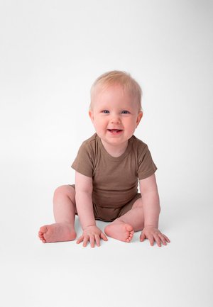 Smilende baby med lyst hår iført en brun buksedrakt, sittende på en hvit overflate med hender og føtter flatt på bakken.