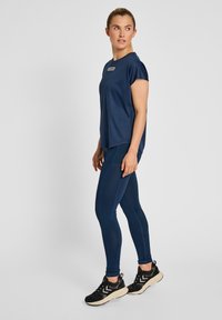 Mörkblå sporttröja med en liten logotyp, korta ärmar, matchas med leggings som har sömdetaljer. Svarta sneakers fulländar outfiten.