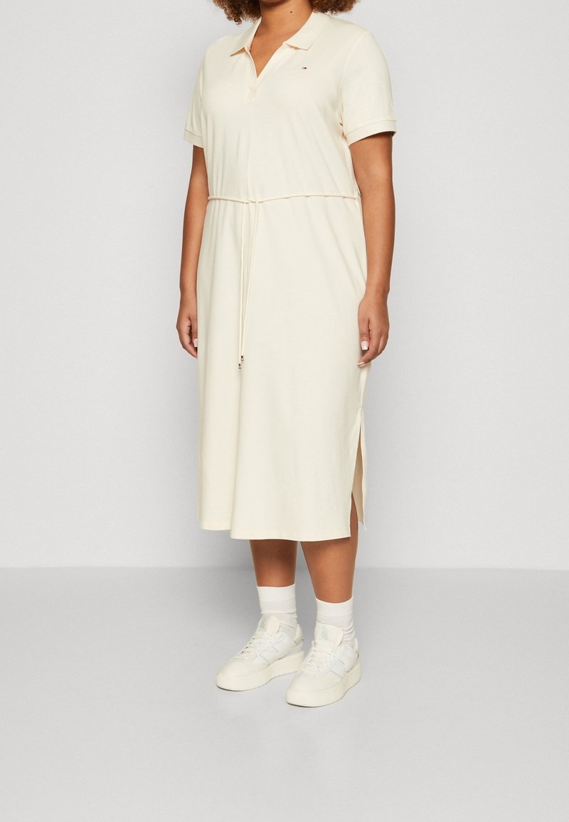 Vestido tipo polo en crema, hecho de una tela suave. Presenta un cuello con solapa, cintura ajustable con cordón, aberturas laterales y se combina con zapatillas blancas y calcetas.