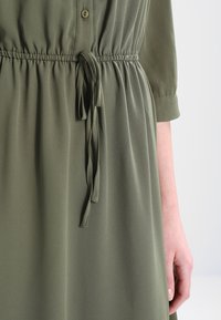 Sukienka w oliwkowym kolorze wykonana z miękkiego, lekkiego materiału; posiada zapięcie na guziki, regulowany pasek w talii oraz podwinięte rękawy. Gładka struktura.