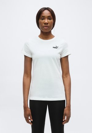 Femme debout, faisant face, portant un t-shirt blanc Puma et un pantalon noir sur un fond clair uni.
