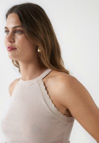 Top sin mangas de color beige claro con detalles de escallos texturizados en los hombros. Material suave con una forma ajustada. Pendientes de aro dorados.