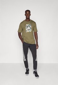 Mand står med hænderne i lommerne iført olivengrønt grafisk t-shirt, sorte træningsbukser med hvide striber og sorte sneakers mod en ensfarvet baggrund.