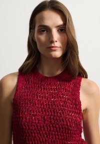 Top senza maniche rosso con un design a maglia aperta e testurizzata. Il collo è rotondo e il tessuto sembra leggero e traspirante.