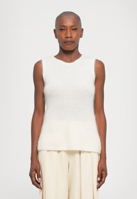 Žmogus su nuskusta galva, apsirengęs be rankovių baltu pūkuotu megztiniu ir kreminėmis plačiomis kelnėmis, stovi priešais šviesiai balta fono.