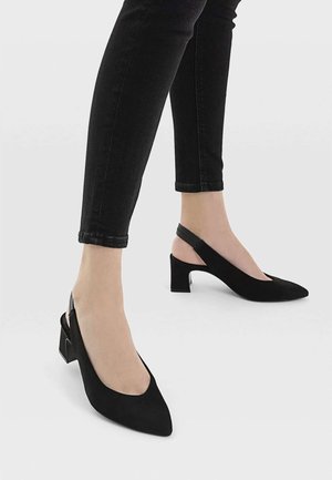 Person iført sorte slingback sko med blokhæl og spidse tæer sammen med sorte, tætsiddende bukser mod en ensfarvet baggrund.