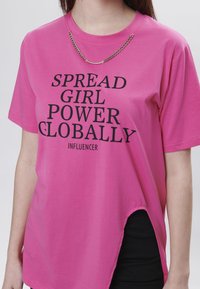 Camiseta de algodón rosa con mangas cortas y un dobladillo irregular. Texto negro que dice "DISEMINA EL PODER FEMENINO A NIVEL GLOBAL" y "INFLUENCER". Detalle de cadena en el escote.