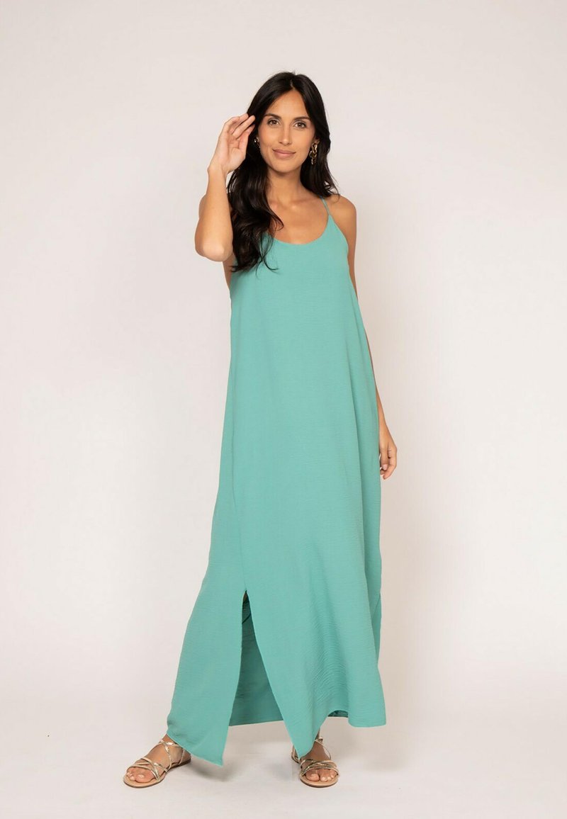 Une longue robe sans manches couleur sarcelle avec une silhouette décontractée, présentant une fente à l'avant et une texture lisse, complétée par des sandales plates métalliques.