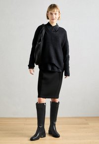 Pull en maille noir à manches larges, superposé à une chemise noire, associé à une jupe noire ajustée et des bottes en cuir noires montantes à talons blocs.