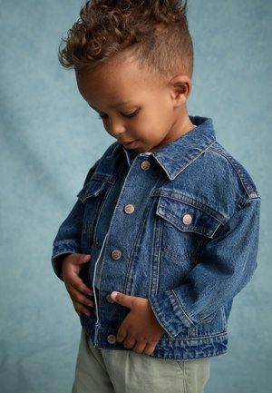 Junges Kind mit lockigem Haar, das eine Jeansjacke und helle Hosen trägt, schaut nach unten, während es die Vorderkanten der Jacke hält.