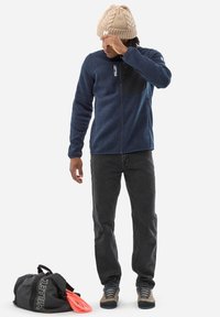 Veste en polaire bleue avec un panneau noir, fermeture éclair à l'avant et col haut. Portée avec un jean gris et un bonnet en tricot beige. Un sac noir avec une corde d'escalade à proximité.