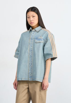 A young woman poses confidently, wearing a light blue denim shirt with orange stripes and beige trousers, against a plain white background.