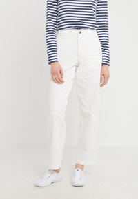 Person som bär vita högmidjade raka byxor, vita sneakers och en marinblå och vit randig långärmad skjorta, som står mot en vit bakgrund.