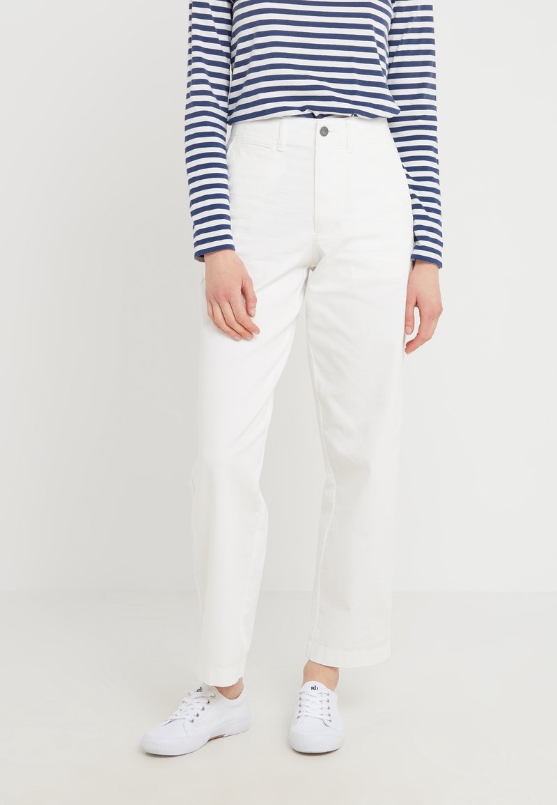 Person som bär vita högmidjade raka byxor, vita sneakers och en marinblå och vit randig långärmad skjorta, som står mot en vit bakgrund.