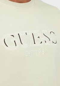Tuvumā attēls ar bēša audumu, uz kura krūtīs ir izšūts "GUESS" logotips un teksts "EST. 1981".