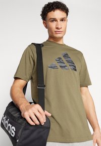 Maslinasto zelena majica s kamuflirajućim crnim Adidas logotipom. Model drži crnu mrežastu torbu s bijelim Adidas brendiranjem.
