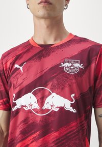 Maglietta sportiva rossa a maniche corte con un motivo di pennellate, caratterizzata da una grafica di un toro bianco e un logo sul petto. Realizzata in tessuto traspirante.