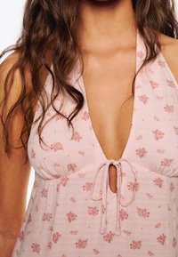Vestido floral rosa claro con un profundo escote en V, detalle de lazo en la parte frontal y tejido texturizado que presenta pequeños patrones de rosas en toda la prenda.