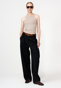 Naine, kellel on seljas beež ilma varrukateta topp, mustad laiad püksid pruuni vööga, pruuni mokassiinid ja tumepruunid prillid, seisab valge tausta ees.