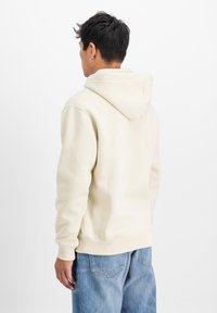 Bēša kapucēta džemperis ar ribotām aprocēm un apakšmalu, ar mīkstu tekstūru. Valkāts ar gaiši ziliem džinsiem, skats no aizmugures.