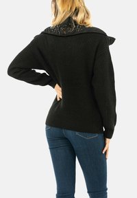 Maglione nero lavorato a maglia con finitura testurizzata e sovrapposizione nera con colletto decorata con borchie. Vestibilità ampia e maniche lunghe, abbinato a jeans blu.