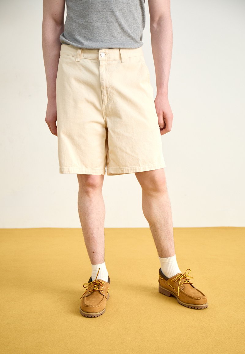 Beige bomullshorts med sidelommer, knappelukking og en avslappet passform, sammen med brune mokasinlignende sko og hvite sokker.