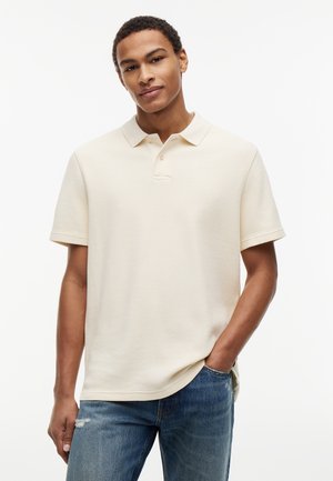 Νέος άνδρας με μπεζ κοντομάνικο πόλο και μπλε τζιν, όρθιος με το ένα χέρι στην τσέπη, μπροστά σε μια απλή λευκή επιφάνεια.