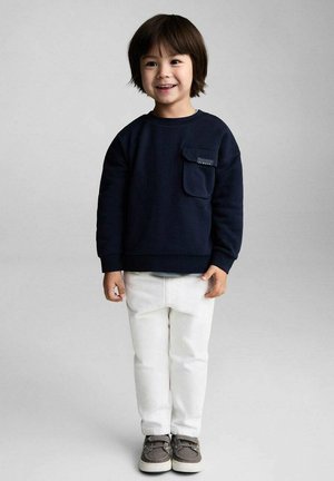 Marinblå sweatshirt med bröstficka, vita jeans och grå sneakers. Barnet har kort, mörkt hår och ler. Enkel, avslappnad outfit.