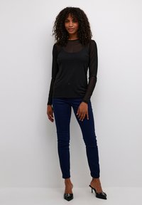 Un haut noir à manches longues avec un panneau avant transparent, associé à un jean skinny bleu foncé et des mules à talons noirs, sur un fond clair.