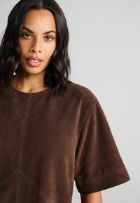 Haut en suède marron à manches courtes, décolleté rond et détails de couture subtils. Texture lisse avec un design épuré et minimaliste.