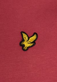 Rød tekstureret stof prydes af et markant gult broderet ørne-logo, omkranset af sort. Designet er simpelt med fokus på emblemets udtryk.