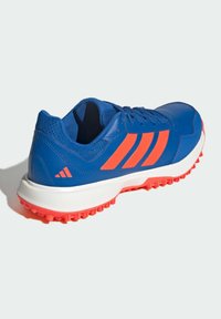 Chaussure de sport bleue avec des bandes orange, semelle blanche et semelle orange, vue de dos sur un fond clair.