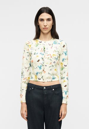 Femme aux longs cheveux noirs portant un chemisier ajusté à fleurs à manches longues boutonné et un jean taille haute foncé, sur un fond uni.
