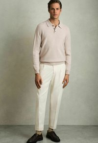 Polo en laine à manches longues rose clair avec un col et une patte de boutonnage à trois boutons, associé à un pantalon blanc et des chaussures marron, sur fond neutre.