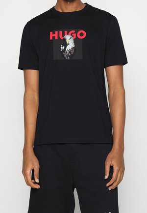 Czarny bawełniany t-shirt z grafiką białogłowego orła i nazwą marki "HUGO" w wyraźnych czerwonych literach. Krótkie rękawy, luźny krój.