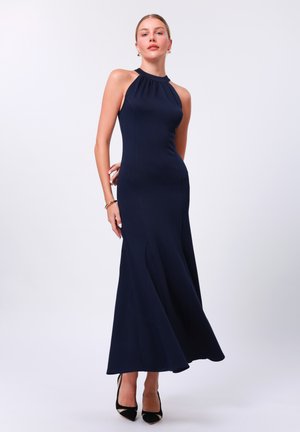 Navy blue halter neck dress, form-fitting with a flared skirt, made of smooth fabric. Black heels with a metallic accent complete the outfit.