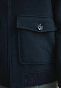 Tummansinisen laivastonsinisen takin tasku, jossa on läppä ja nelireikäinen nappi, sekä alareunassa resoripäälli.