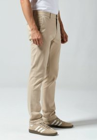 Beige Chinos in schlanker Passform mit Reißverschluss, seitlichen Taschen und glatter Textur, kombiniert mit hellbraunen Sneakern und weißen Streifen.