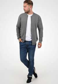 Siva zip-up kardigan z ribanim vzorom, oblečen nad belo srajco, v kombinaciji z modrimi kavbojkami in črnimi supergami. Model pozira od strani.