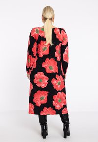Robe noire avec un grand imprimé floral rouge, manches longues et longueur mi-mollet, associée à des bottes à talons noirs brillantes.