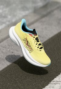 Adidași de alergare de un galben strălucitor, cu un material superior respirabil, șireturi negre și logo-ul HOKA. Prezintă o talpă albă cu amortizare, având un accent de albastru deschis.