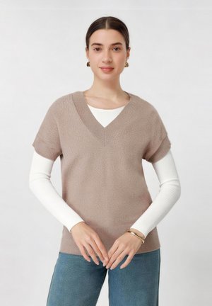 Ženska s spetimi lasmi nosi bež V-izrez kratko majico z belimi dolgimi rokavi, modre kavbojke, zlate uhane in zapestnico, roke ima sklenjene.